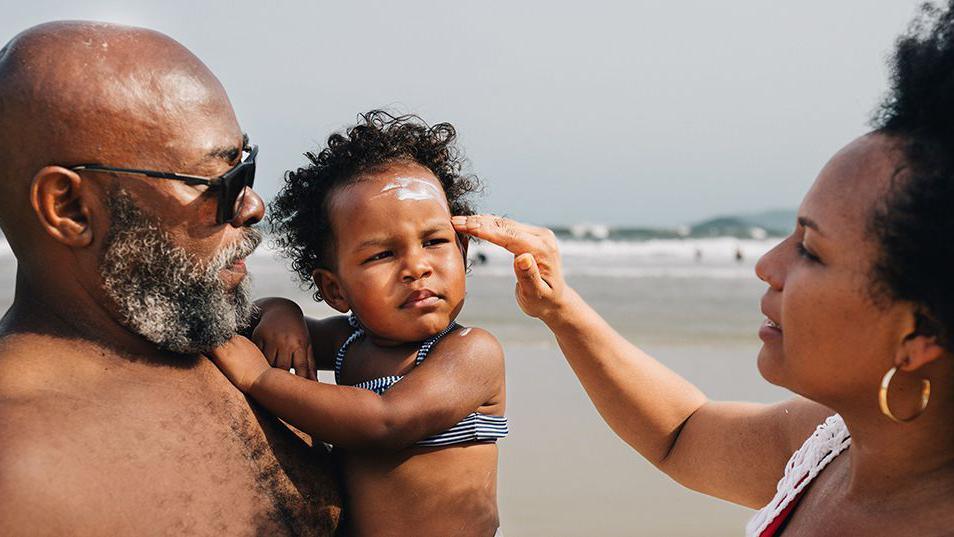 On a beach, a mum applies sunscreen to her young daughter who is in her father's arms.