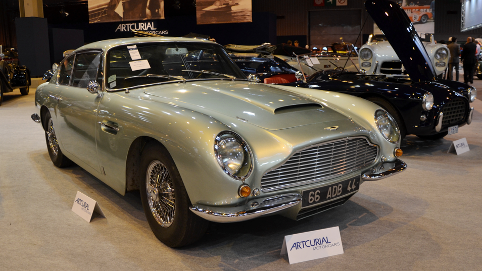 Silver-grey classic Aston Martin-style coupe displayed at an indoor collector car show, viewed from the front-left angle. The car features a large chrome grille, round headlights, wire-spoke wheels, and polished chrome trim. Display signs reading “ARTCURIAL MOTORCARS” sit in front of the vehicle, while other vintage cars and exhibition displays are visible in the background under bright showroom lighting.