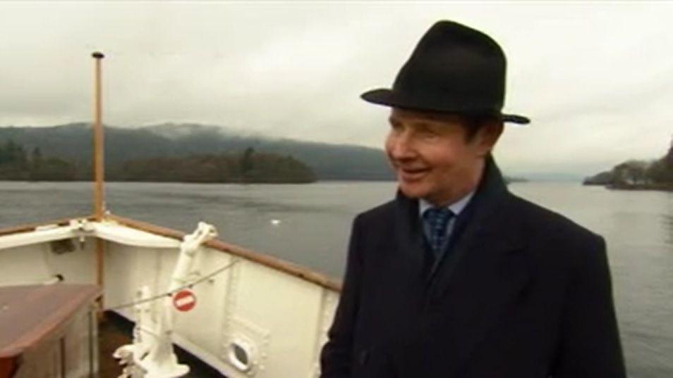 Jonathan Denby pictured standing on a boat in the Lake District in March 2018. He is wearing a smart black hat and black overcoat, with a large lake and wooded hills behind him.