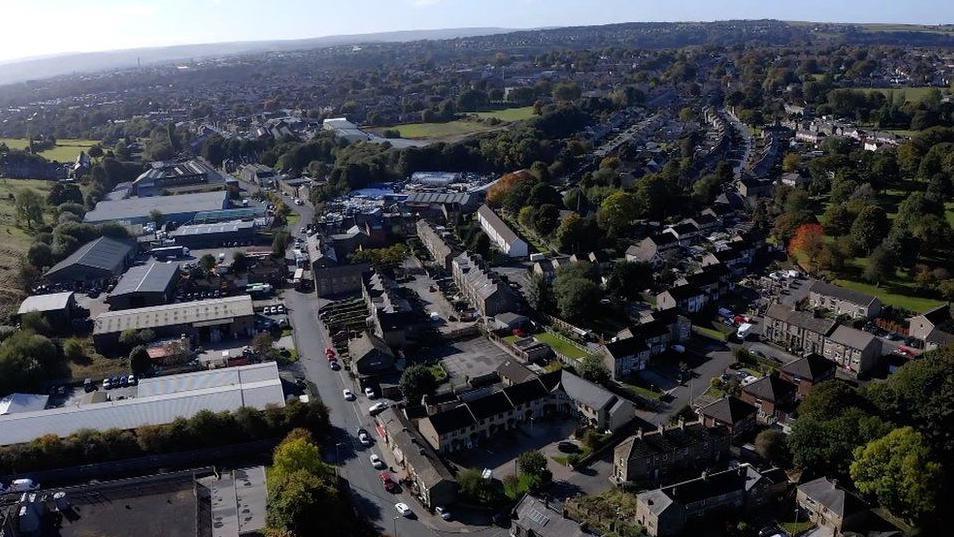 Aerial view of the village of Holmfield. It shows a mixture of industrial units located near houses with hills and trees surrounding the landscape. 