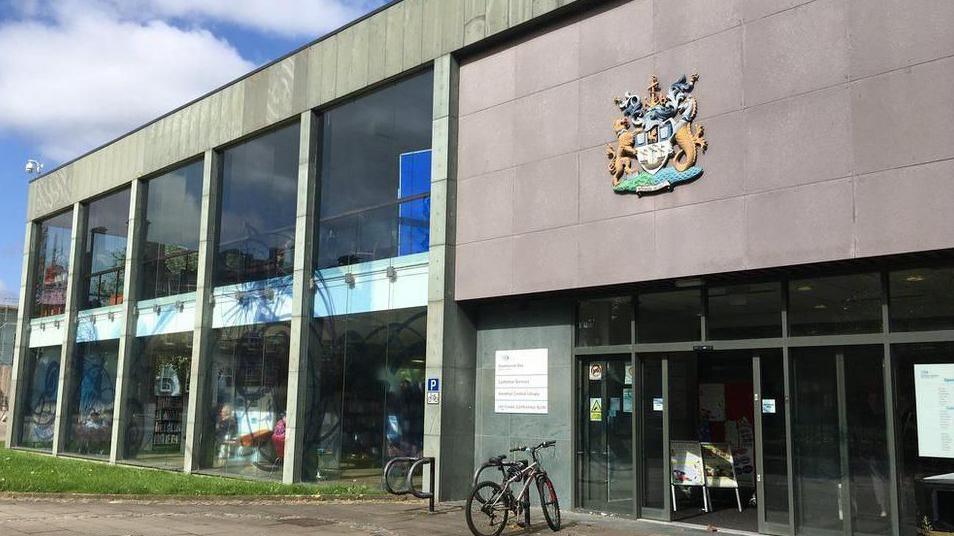 View of the entrance to Stockton Council's offices. To the right there are glass doors, with a council crest on the wall above them, and a parked bicycle outside. The left side of the two storey building is mainly glass.