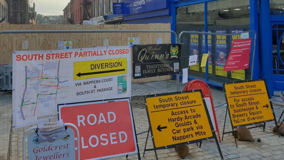 Multiple signs in front of a wooden fence that's sealing off part of the shopping street. One has a map and reads South Street partially closed, diversion. Another says road closed. Others mention specific businesses and have arrows to direct people for access.