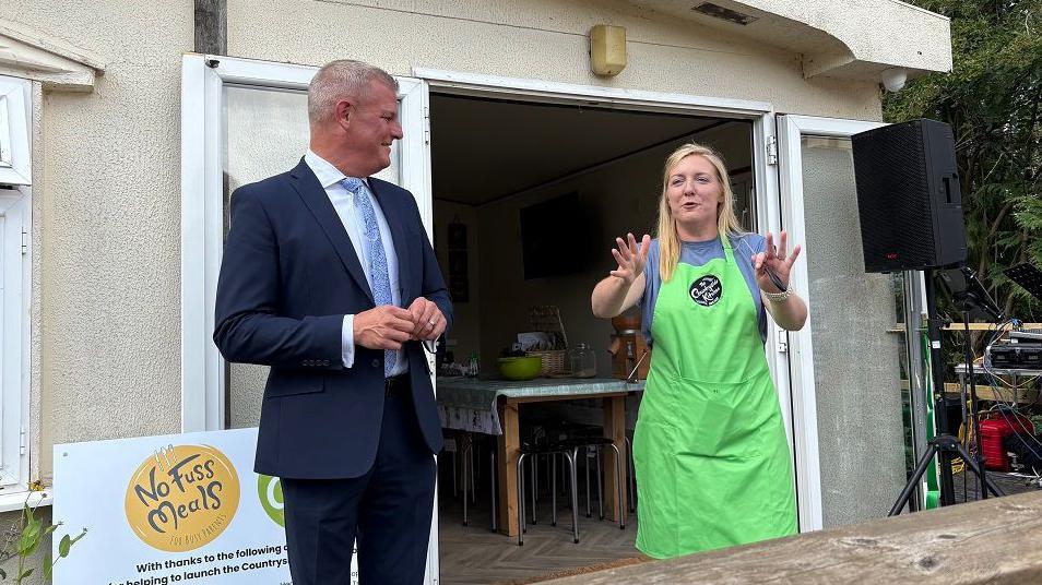 Stuart Andrew with short white hair, wearing a dark blue suit and light blue tie. Milly Fyfe has long blonde hair and is wearing a green apron over a blue T-shirt. She has her hands outstretched while talking. They are standing outside a single-storey wooden classroom with doors open revealing a table inside.