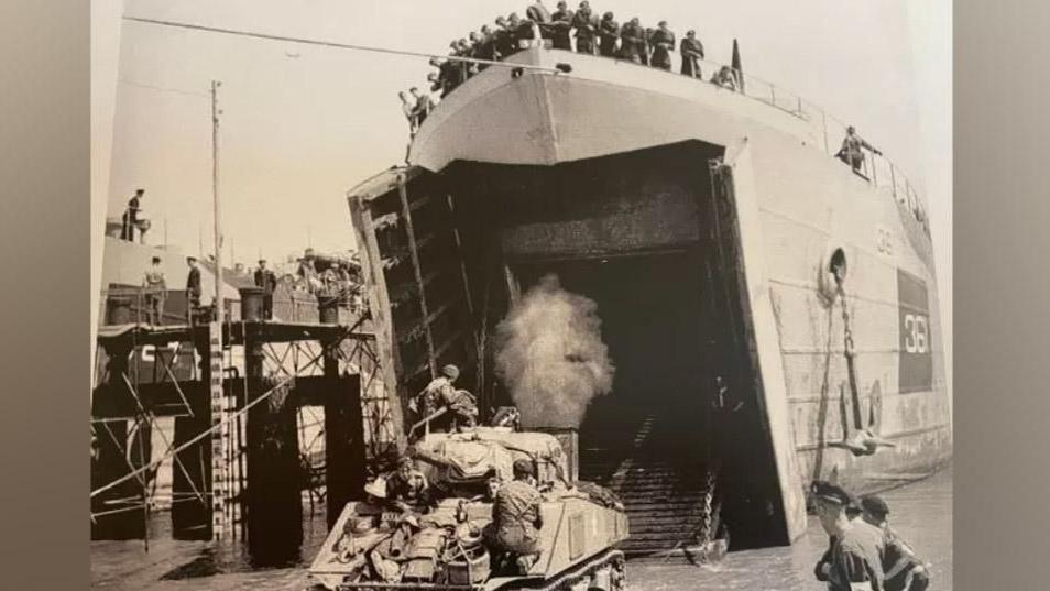 A black and white image of a tank leaving a ship during the D-Day landings. You can see men standing at the top watching the tanks come off.