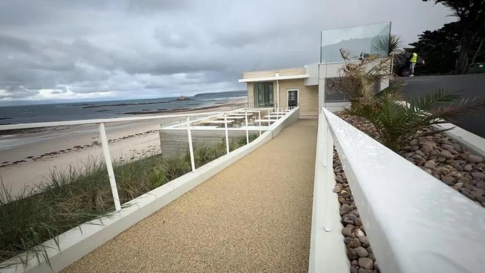 A pathway leads down to a beachside building with tables seen outside and to the left a stretch of white sand bordered by the sea