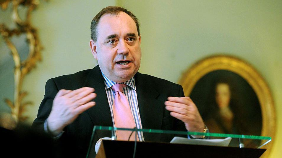 Alex Salmond speaking in Bute House in 2008. He is standing at a glass podium, wearing a black suit, checked shirt and pink tie. A portait in an oval gold frame hangs on the wall behind him.