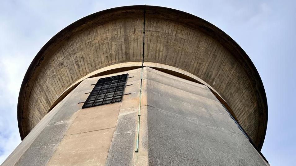 A close-up shot of the water tower, with the camera pointing up at the dome. Concrete walls rise up to the dome looming above.