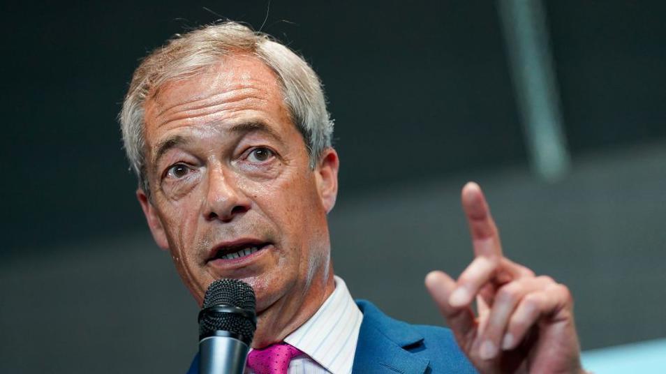 A head and shoulders picture of Mr Farage. He is wearing a blue suit and pink tie and is holding a microphone. The background is grey and blurred.