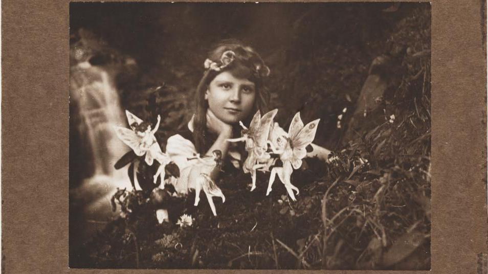 Sepia photo of young girl resting her chin on her hand gazing at a collection of small fairies with wings in the foreground ahead of her in a garden