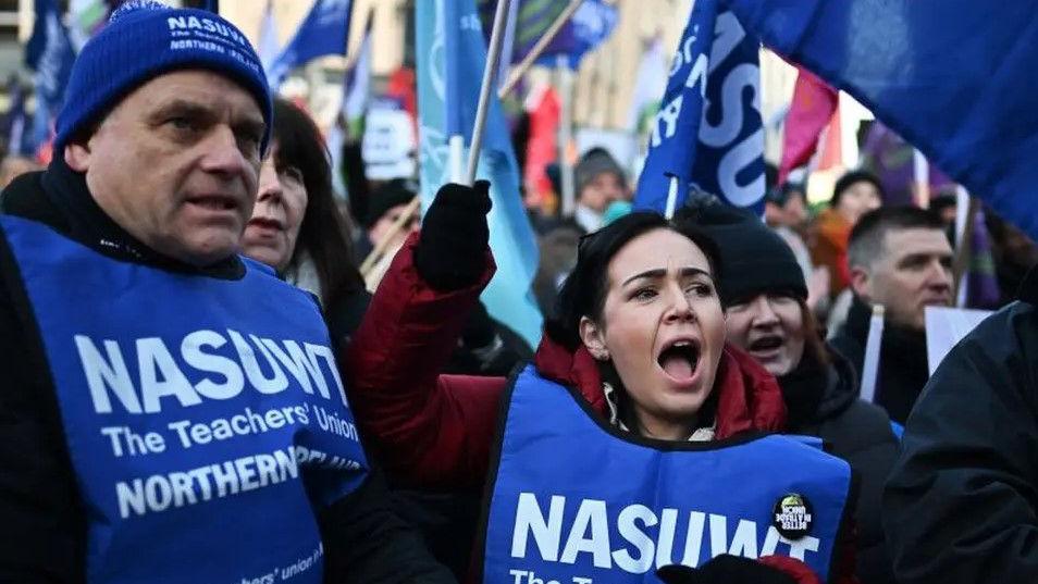 Teachers on strike, protesting with flags. A man on the left wears a blue NASUWT vest and hat. A woman to his right wears an identical blue vest. He is wearing a black hoody and she is wearing a gret hoody. She is holding a blue NASUWT flag. There is a crowd behind them, with people holding flags and placards.