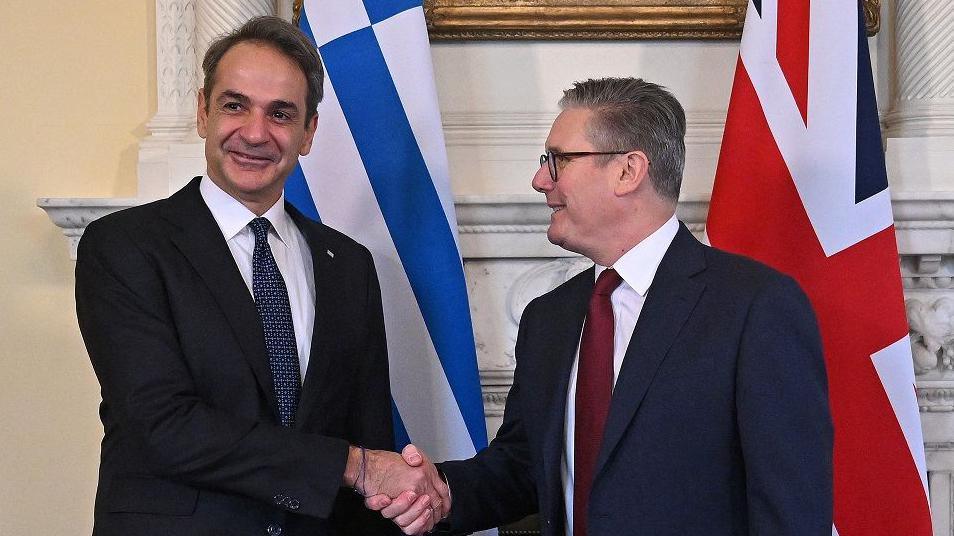 Starmer and Mitsotakis smile and shake hands in No 10. Starmer is looking at Mitsotakis, while Mitsotakis is looking at the camera. Their countries' flags are behind them