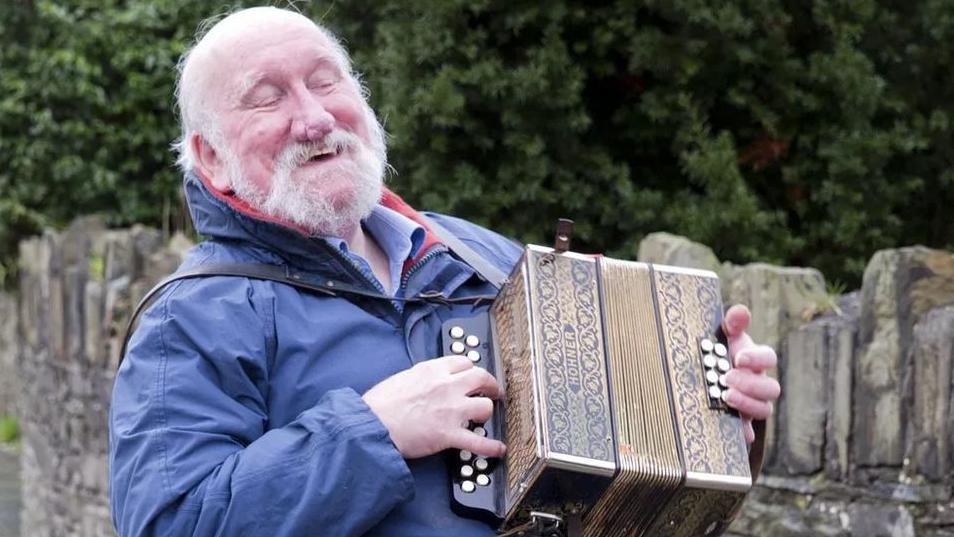 John Kaneen playing an accordion. He is wearing a blue coat and is smiling. There is a stone wall behind him along with green trees.