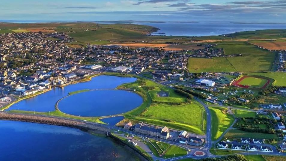 An aerial view of Orkney by the sea which shows a green landscape with homes and buildings connected by roads, large blue lakes and a running track.