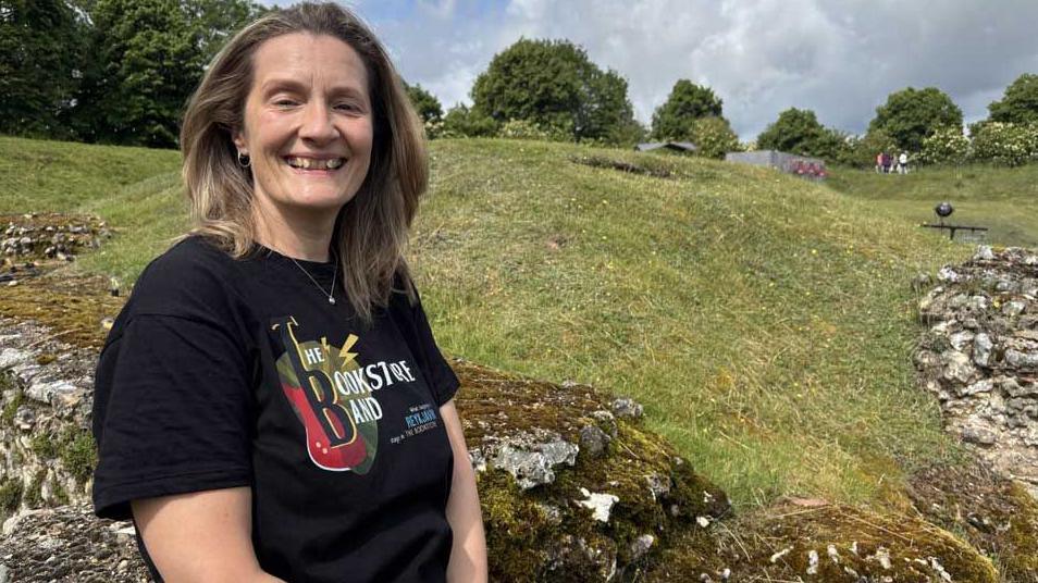 Emma Wright is standing in the ruins of the Roman Theatre at Verulamium and you can see flint walls behind her. She has shoulder-length dark blonde hair and is smiling at the camera. She wears a black T-shirt with white writing on it.