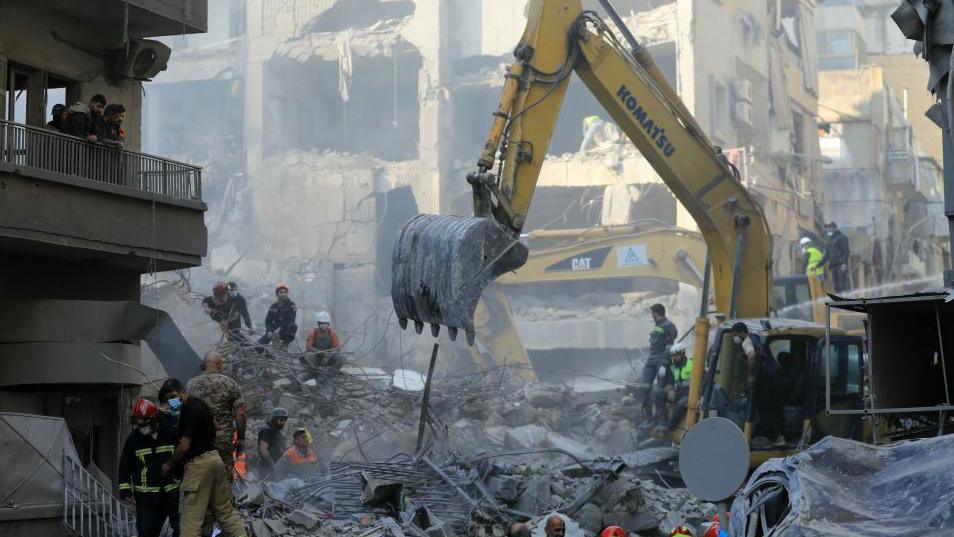 The scene of destruction following a massive Israeli strike on central Beirut. An excavator is digging through the rubble as dozens of rescue workers move around. It's daylight and dust hangs in the air.