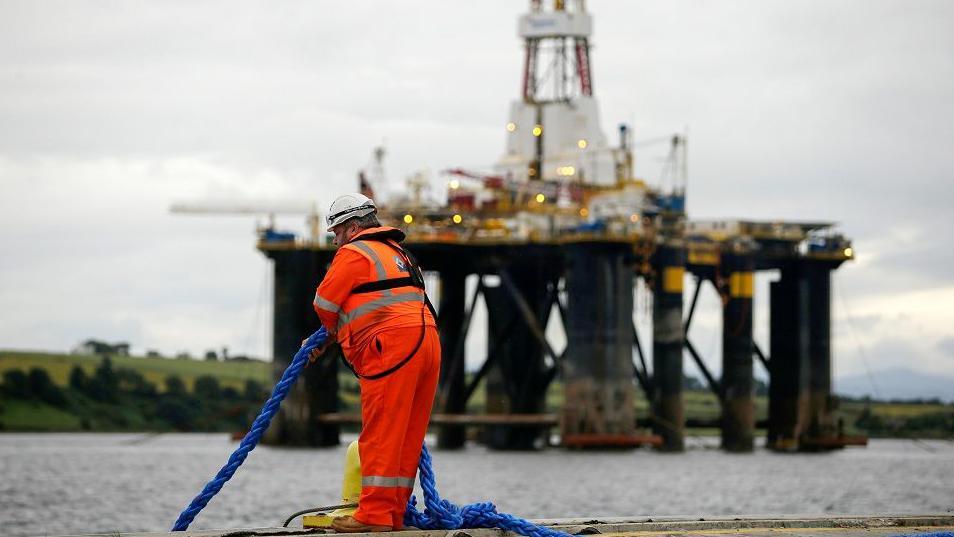 An employee in an orange high vis suit stands on the quay side as he pulls in a rope in view of a mobile offshore drilling unit in the Port of Cromarty Firth in Cromarty