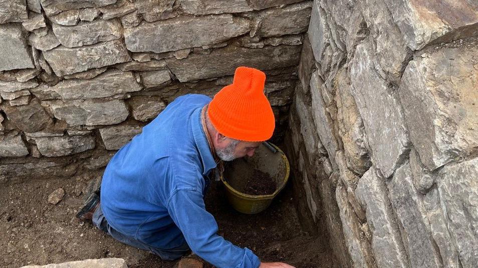 An archaeologist in a denim shirt and bright orange beanie is kneeling at the site of the medieval toilet. There is a bucket next to him and he is surrounded by a stone wall.