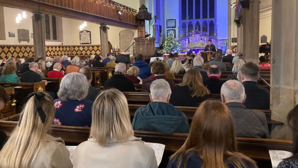 We see the back of the heads of people in wooden pews inside the minster, with stone pillars going up towards the head. Many of them wear coats and jackets, the nearest are women with long hair to or past their shoulders.