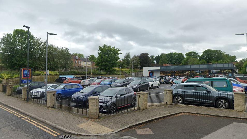 A nearly full car park in front of a single storey Aldi supermarket. Trees in the background.
