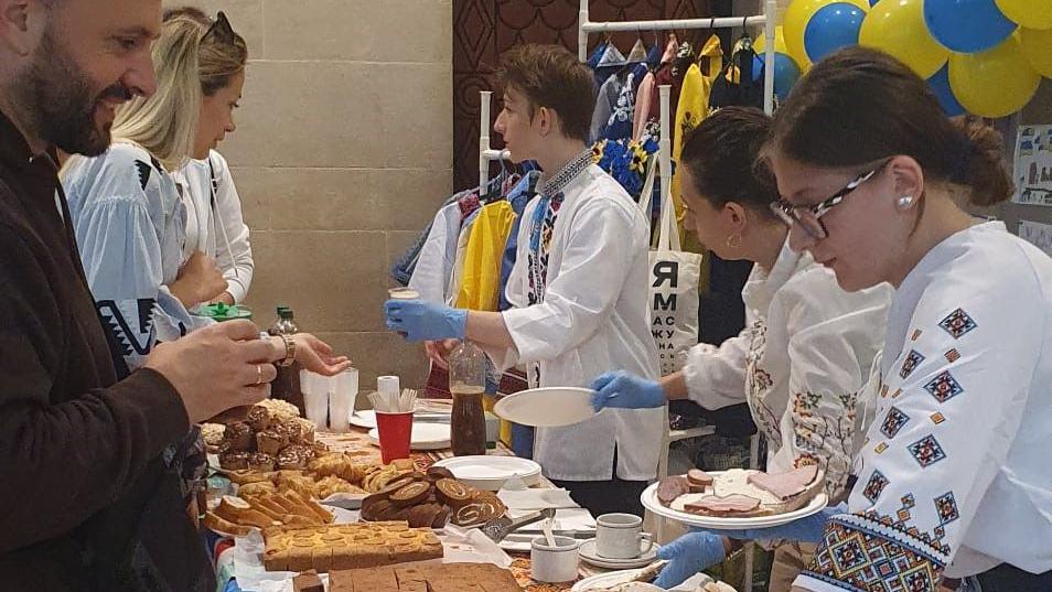 People wearing white outfits serving food to others with yellow and blue balloons behind them