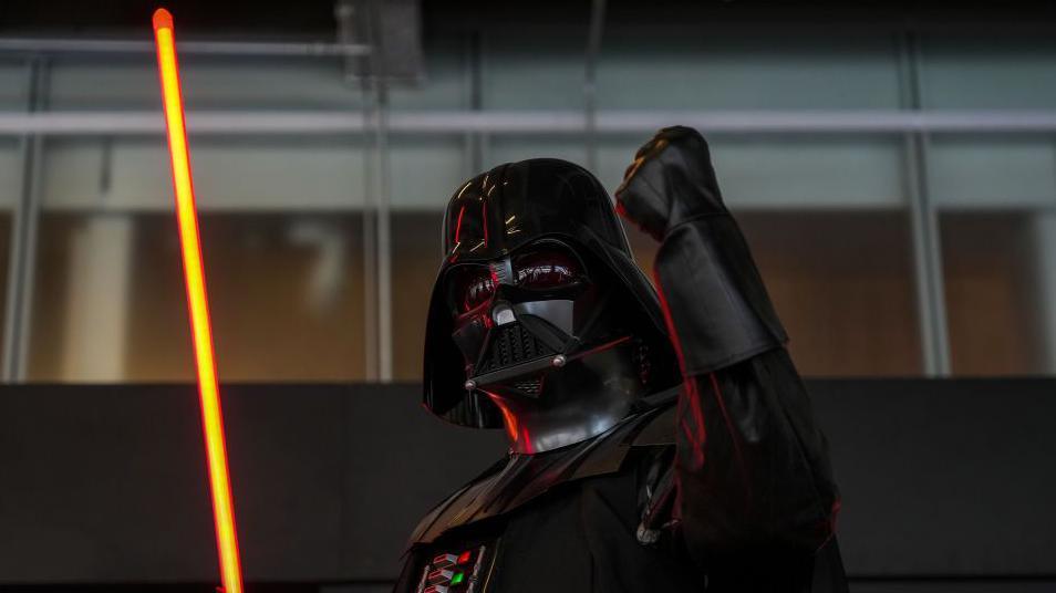 Darth Vader holds a red lighsaber up and has one hand in the air