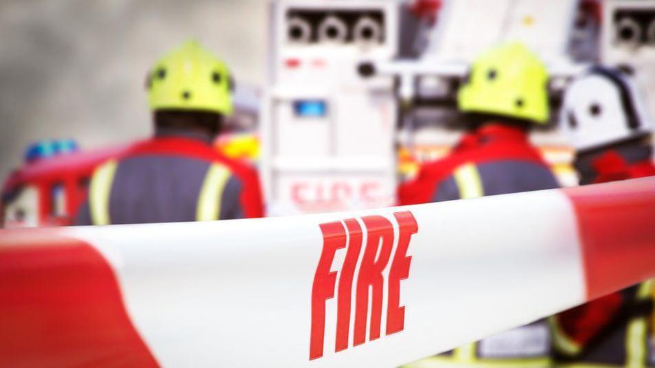 Red and white tape with the word 'fire' written on it. In the background, two firefighters can be seen at the back of a fire truck