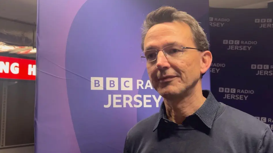 Kirsten Morel - a man wearing rimless rectangle glasses, a navy collared shirt and a navy jumper on top. Behind is the purple BBC Radio Jersey banner.