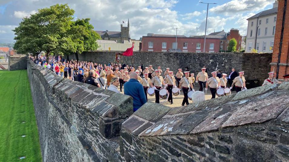 Loyal order members march in uniform, some holding drums and some holding flags, on Londonderry's historic stone walls