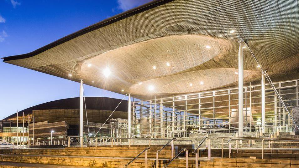 A brightly lit exterior picture of the front of the Senedd building with the Wales Millennium Centre in the background