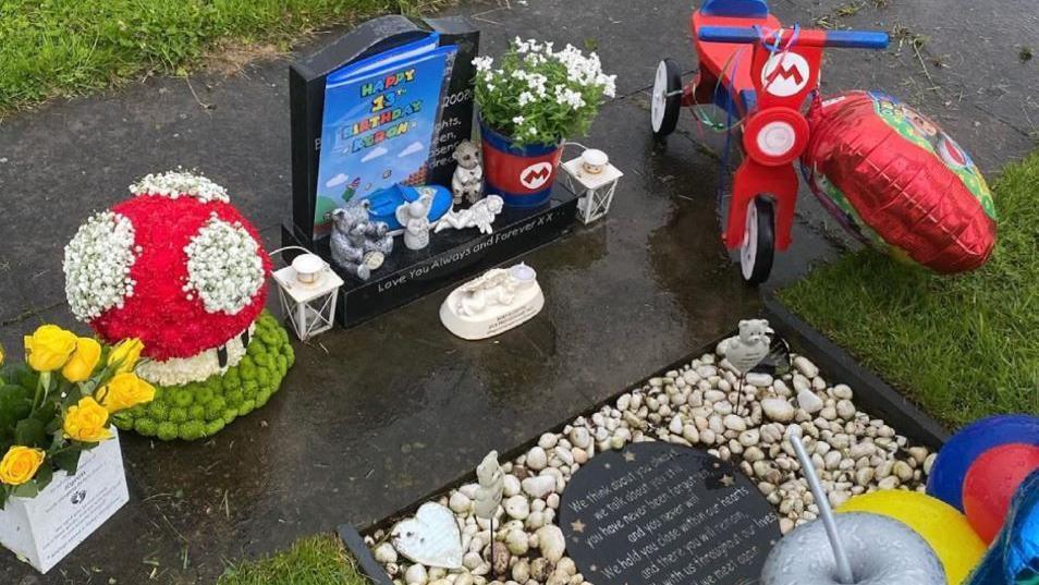 A child's grave, with supermario decorations around it including a floral mushroom decoration, red plant pot with an M motif and a mini red scooter. There are also yellow roses in a box and ornaments at the foot of a small black headstone. A flat plaque lies in front of the headstone, on a bed of stones. Grass surrounds the plot.