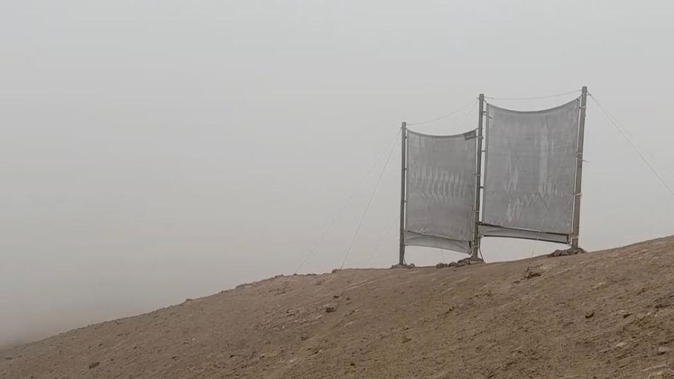 An experimental fog harvesting system - consisting of two sheets of fine mesh nets, each suspended between two poles. The nets sit on a barren hill in the mist