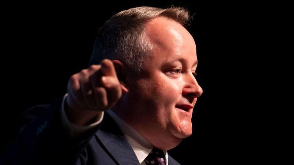 Darren Millar is pointing towards the camera while looking away at a right angle towards an audience. He is wearing a suit, tie and shirt, and is stood before a dark background.