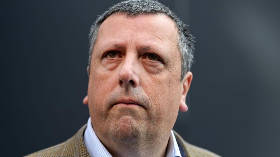 A man with short, greying hair looks towards the frame with a focused facial expression. He is wearing a white patterned shirt and light brown jacket.