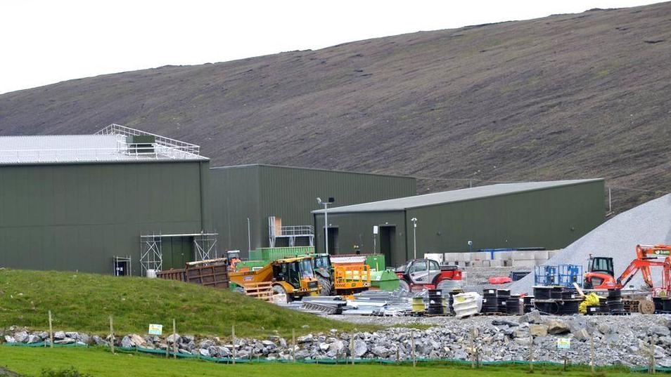 Three corrugated metal-walled buildings dominate the site. There is a yard with construction vehicles, including a tipper truck and excavators.  