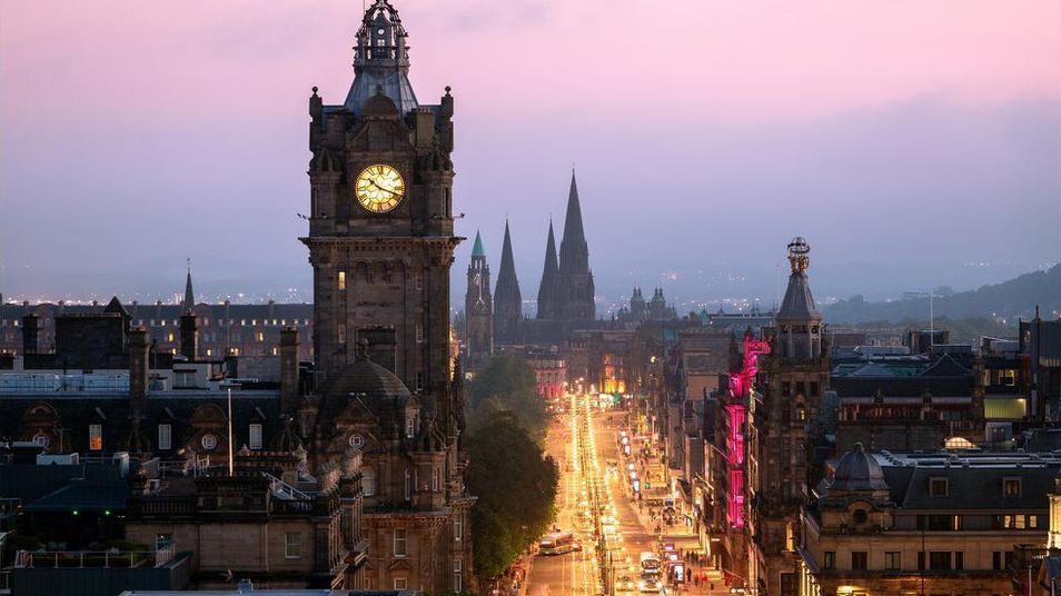 The Balmoral Hotel clock rises into a purple night sky next to a lit-up  Princes Street, glowing golden from the street lights. We can see church spires at the far end and other buildings line the other side of the street. The picture was taken from height so we are looking down on the street.