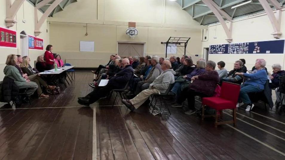 A community hall with about 50 people sitting in rows facing a panel of people where a woman is standing up and talking