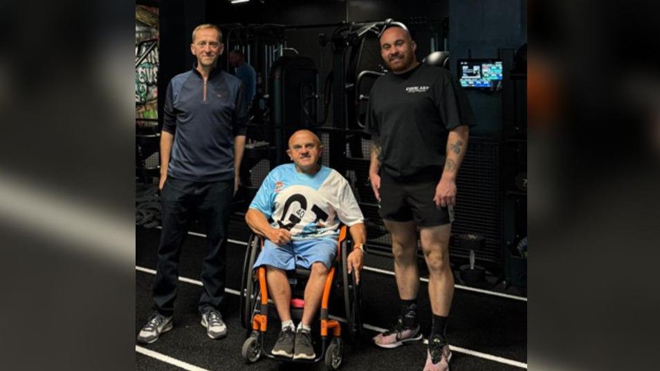 Steve Matthews, chairman of the Archie Matthew's Trust, wearing a navy sports top, black sports trousers and white and grey trainers, standing with his hands in his pockets. Les Hampton is in his wheelchair wearing blue shorts and a blue and white t shirt. He has dark grey trainers and is smiling at the camera. Gym manager Jo Allen is wearing a black t shirt and black shorts and a pair of light coloured trainers. There are some kind of TV screens in the background.