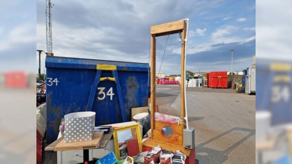 A wooden guillotine, with an electric motor and a blade splatted with fake blood, stands among a pile of rubbish at Grimsby waste recycling centre. A blue skip, for holding waste, stands to the left.