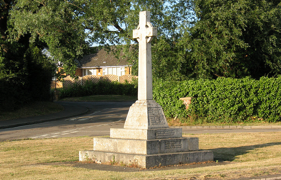 War memorial on a lawn, with road junction, hedging, trees and a bungalow in background 