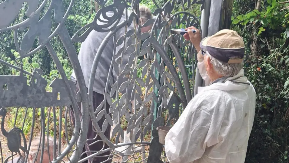 Two men paint and tend to the grey metal gates at the reserve.One man has overalls on and a cap and glasses. He is painting part of the gates.