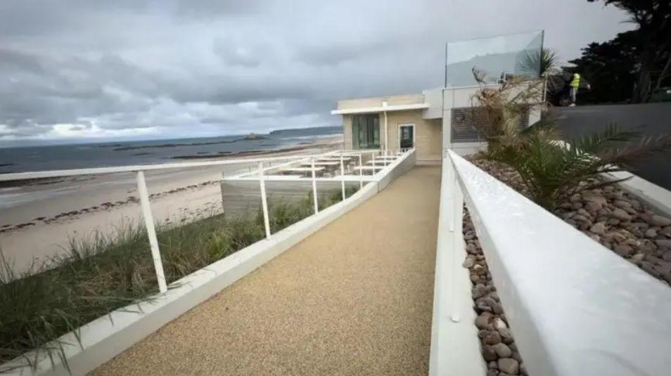 A photo of a modern looking building, with white railings, glass and a long entrace way, on the headland next to St Ouen's beach. The sky is overcast.