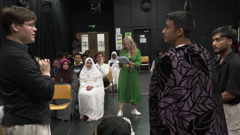 A group of teenage students rehearsing a scene from a Shakespeare play. There are two boys standing in the foreground, one wearing a cape, and several people sitting down watching. A woman in a green dress holds a script watching them. 