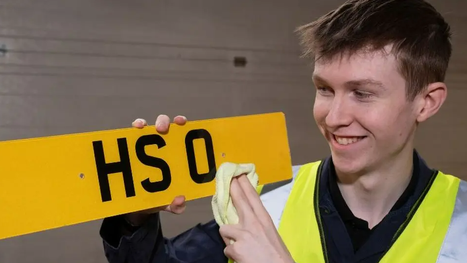 A man polishing the HS 0 number plate. The plate is yellow with black text. The man is young. He has dark hair. He is wearing a dark-coloured top with a bright green hi vis vest over the top.