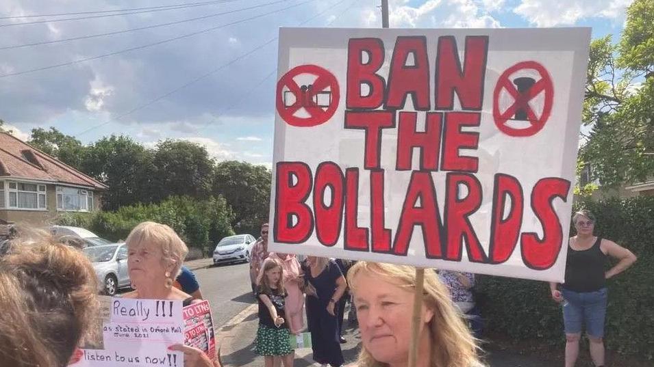 An anti-LTN protest on the side of an Oxford street. A woman with fair hair at the front holds a placard saying 'Ban the Bollards' in red.