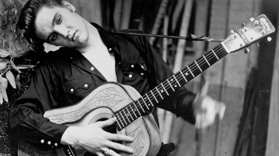 Elvis Presley poses for a portrait holding an acoustic guitar in 1956. He is wearing a dark-coloured shirt and is resting his head against a tree.