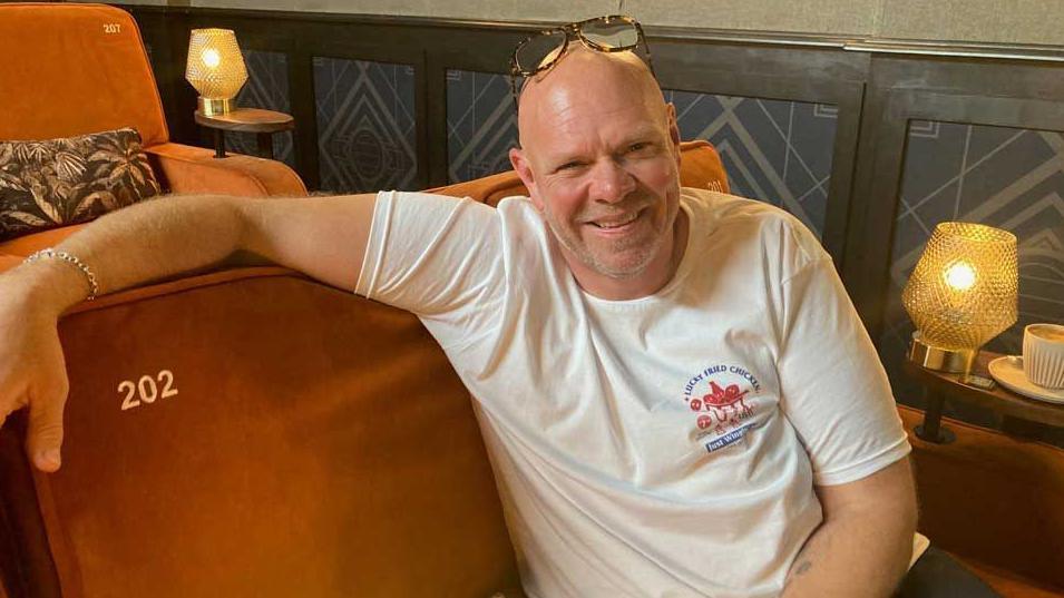 Smiling chef Tom Kerridge wearing a white T-shirt and sitting down on a brown padded armchair