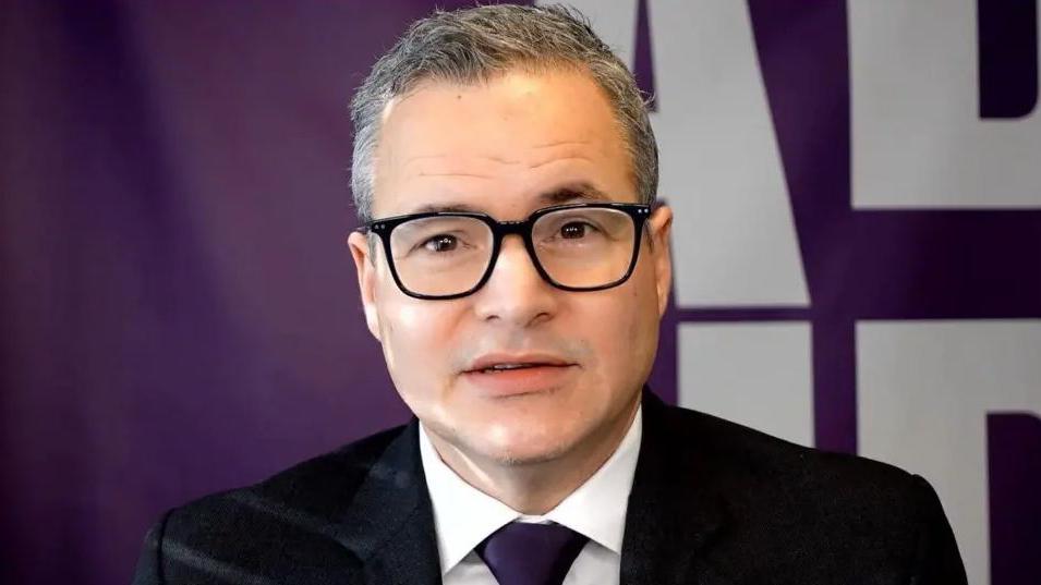 The person is wearing a dark suit jacket with a white shirt and a deep purple tie. The background features a purple banner with large white letters, suggesting a formal or professional setting such as a conference or event.