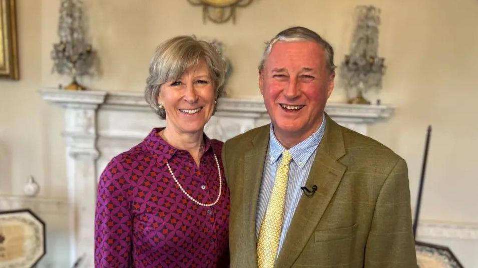 Sir Thomas and Lady Ingilby stand in front of an ornamental fireplace. Lady Ingilby wears a purple shirt and a pearl necklace and earrings. Sir Thomas wears a green tweed suit, blue striped top and yellow tie.