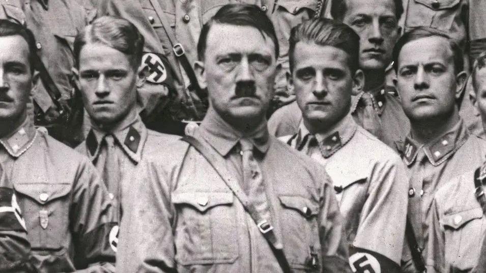 Adolf Hitler staring at camera next to young unsmiling men in Nazi uniform in a black and white photo.