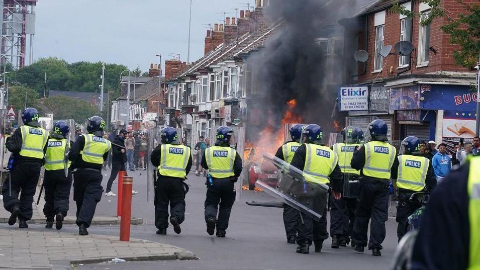 A line of police officers in riot gear approaching a group of rioters. Between the two groups a car is burning in the middle of the high street.